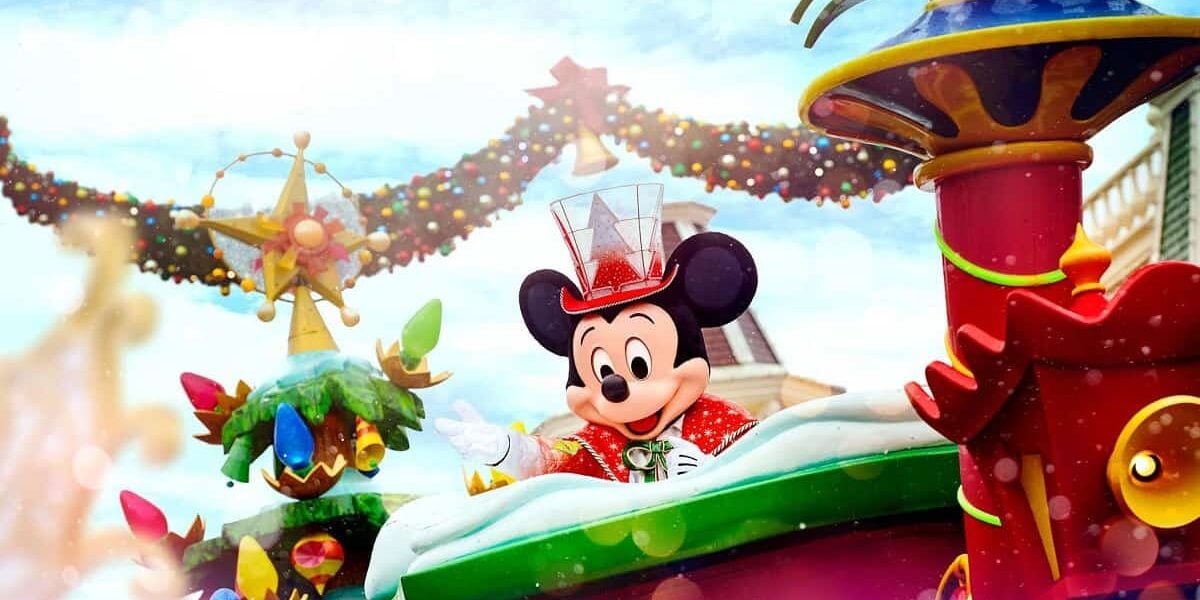 Mickey Mouse in a cheerful Christmas outfit, including a red jacket and a clear top hat, riding on a colorful holiday-themed float decorated with ornaments, garlands, and lights.