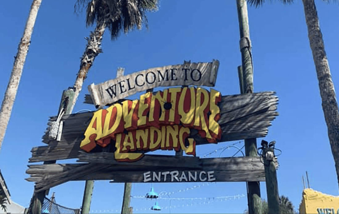 A rustic wooden sign in front of palm trees reads "Welcome to Adventure Landing, Entrance" against a clear blue sky. The vibrant yellow and red lettering stands out against the wooden backdrop, indicating an inviting destination for visitors.