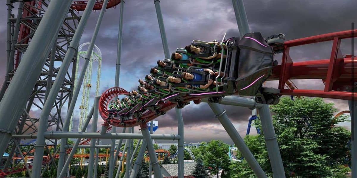 People ride North America's fastest tilt roller coaster at Cedar Point, featuring several loops and twists. The sky is overcast, and the ride's red and grey tracks stand out. Trees and other attractions are visible in the background.