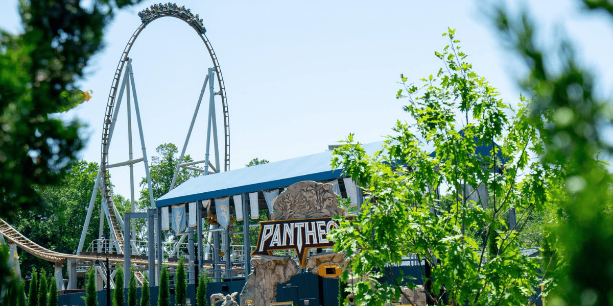 A thrilling roller coaster named Pantheon is visible through some green foliage at Busch Gardens. The ride features a large, vertical loop and tracks that twist and turn. A covered platform for boarding is seen in the foreground with the Pantheon sign prominently displayed.