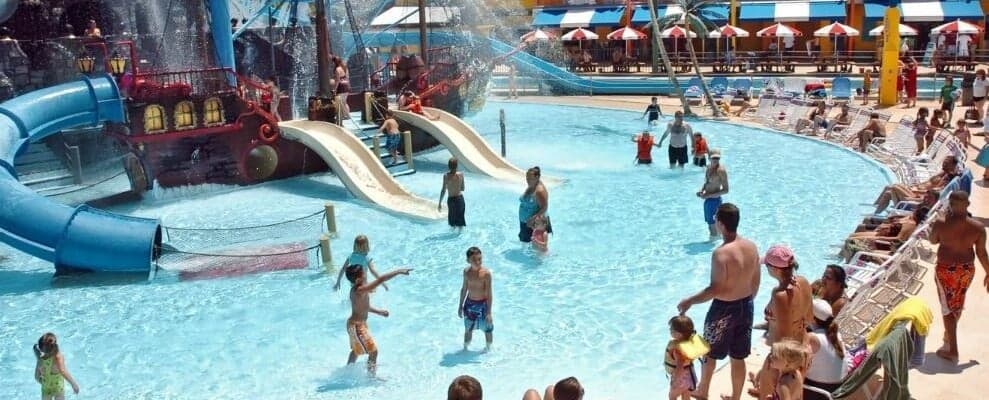 Children and adults enjoy a sunny day at a water park. Kids play in a pool with waterslides and a pirate ship-themed structure, while others relax on lounge chairs nearby. The area is bustling with activity, and colorful umbrellas provide shade in the background.