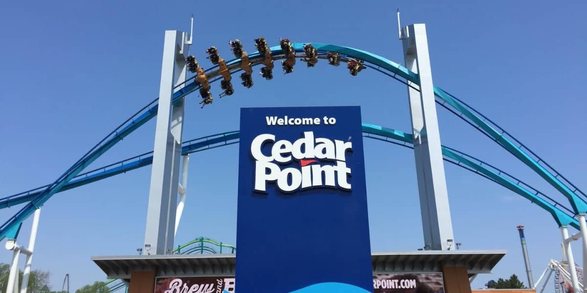 A large blue sign at the entrance of Cedar Point amusement park with the text "Welcome to Cedar Point" in white. Above the sign, people are experiencing an exhilarating roller coaster ride, soaring through the air with the blue track in clear view against a bright sky as PETA advocates nearby.