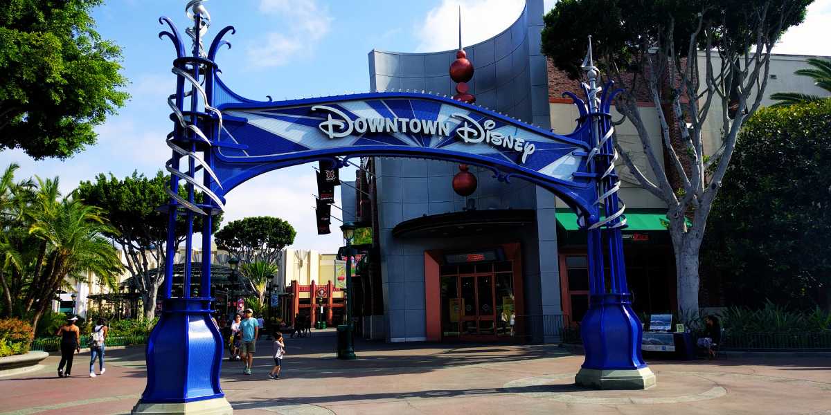 Decorative blue entry arch bearing the "downtown disney" sign above a walkway leading to various shops and eateries, with people walking and green trees under a clear sky.