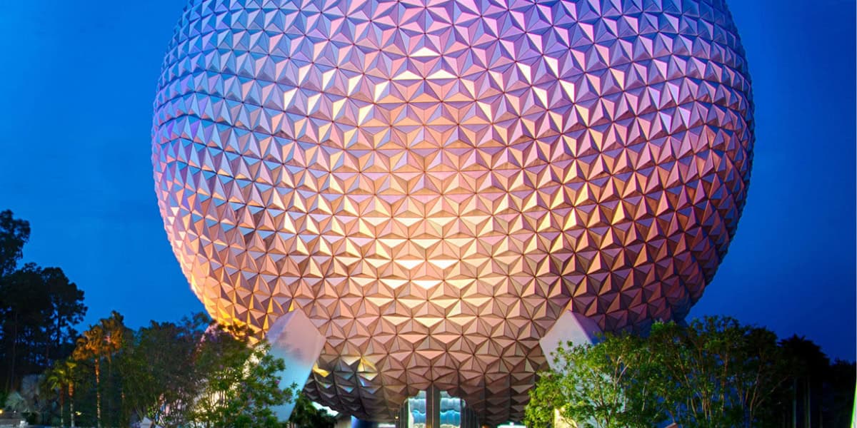 A large geodesic sphere with a triangular pattern, lit up with vibrant blue and purple lights against a twilight sky. The sphere is elevated on angled supports, surrounded by trees and greenery at its base.