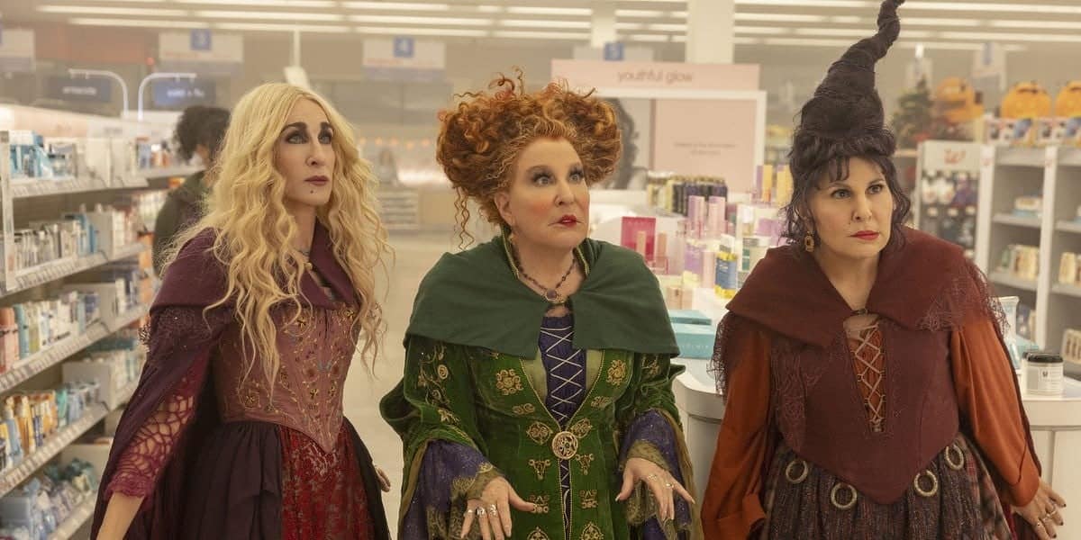 Three women dressed as witches stand in a brightly lit store aisle. They have striking hairstyles and colorful costumes: the first has long blonde hair, the middle has curly red hair, and the third has dark hair styled in an elaborate cone shape. They appear surprised.