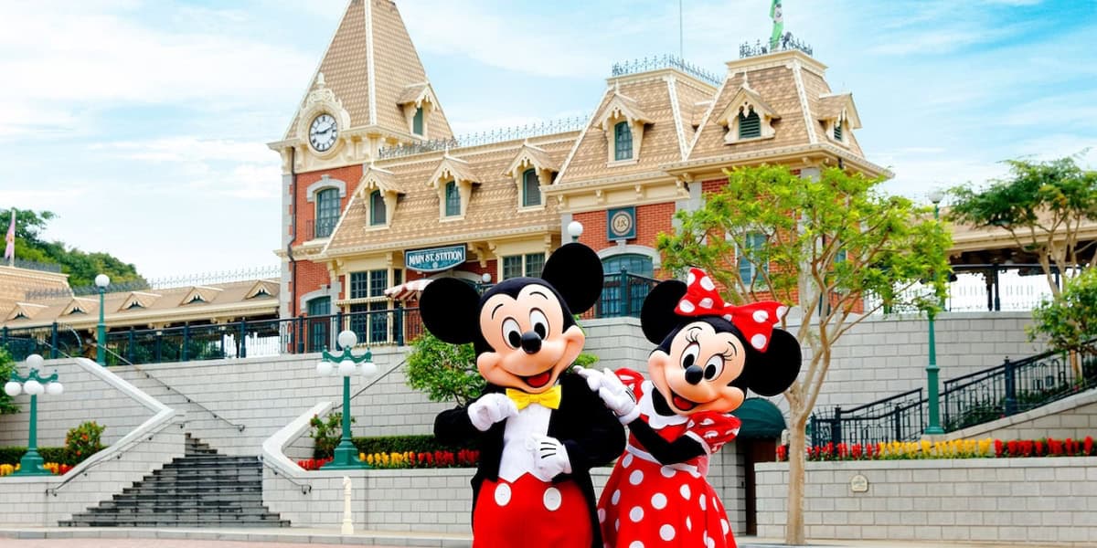 Mickey Mouse and Minnie Mouse, the iconic Disney characters, are standing together in front of a charming station building at Disneyland. Mickey is wearing his classic red shorts and yellow shoes, while Minnie is in her red polka-dot dress and matching bow.