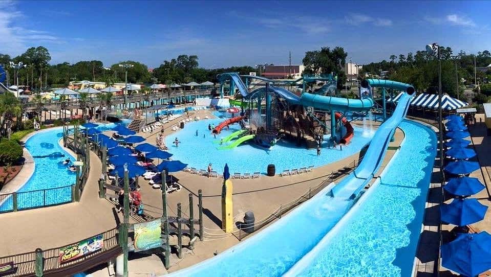 Wide-angle view of a water park featuring multiple slides, including a tall blue one in the foreground, emptying into a large pool. Lounge chairs with blue umbrellas, palm trees, and various colorful attractions are scattered around the area with people enjoying the park.