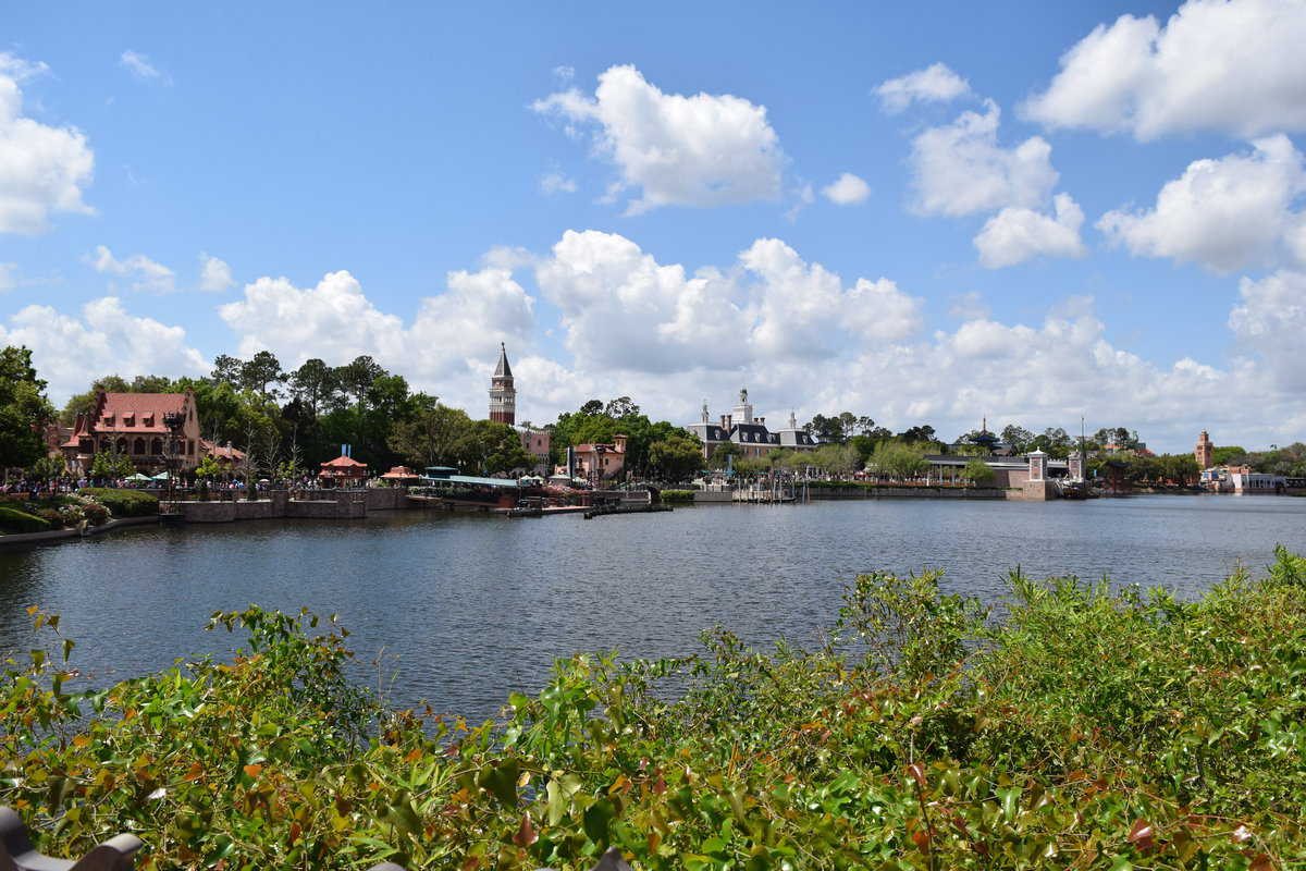 A serene lakeside view with lush greenery in the foreground, a picturesque village with unique architecture along the shore caught in the worst situation, and a backdrop of a clear blue sky dotted with fluffy white