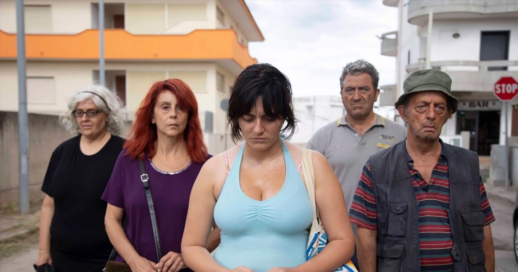 Five people, three women and two men, stand on a street with serious expressions. The woman in front wears a light blue tank top, looking down. In the background, the buildings and stop sign evoke an Avetrana scene—far from Hollywood's glitz.