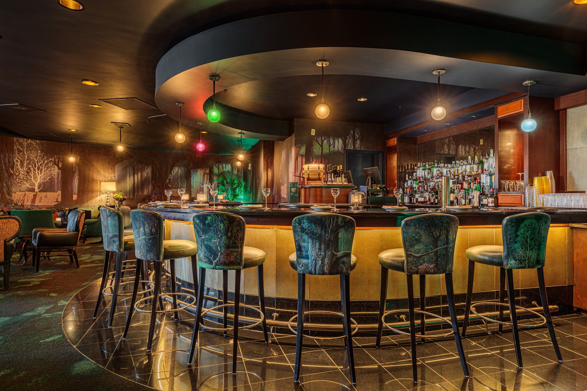 The Broken Spell Lounge at Disneyland is a dimly lit, elegant bar with a sleek circular counter featuring high-backed stools with teal upholstery. Soft lighting creates a cozy ambiance, while the backdrop includes a well-stocked bar and subtle wall art, enhancing the chic atmosphere.