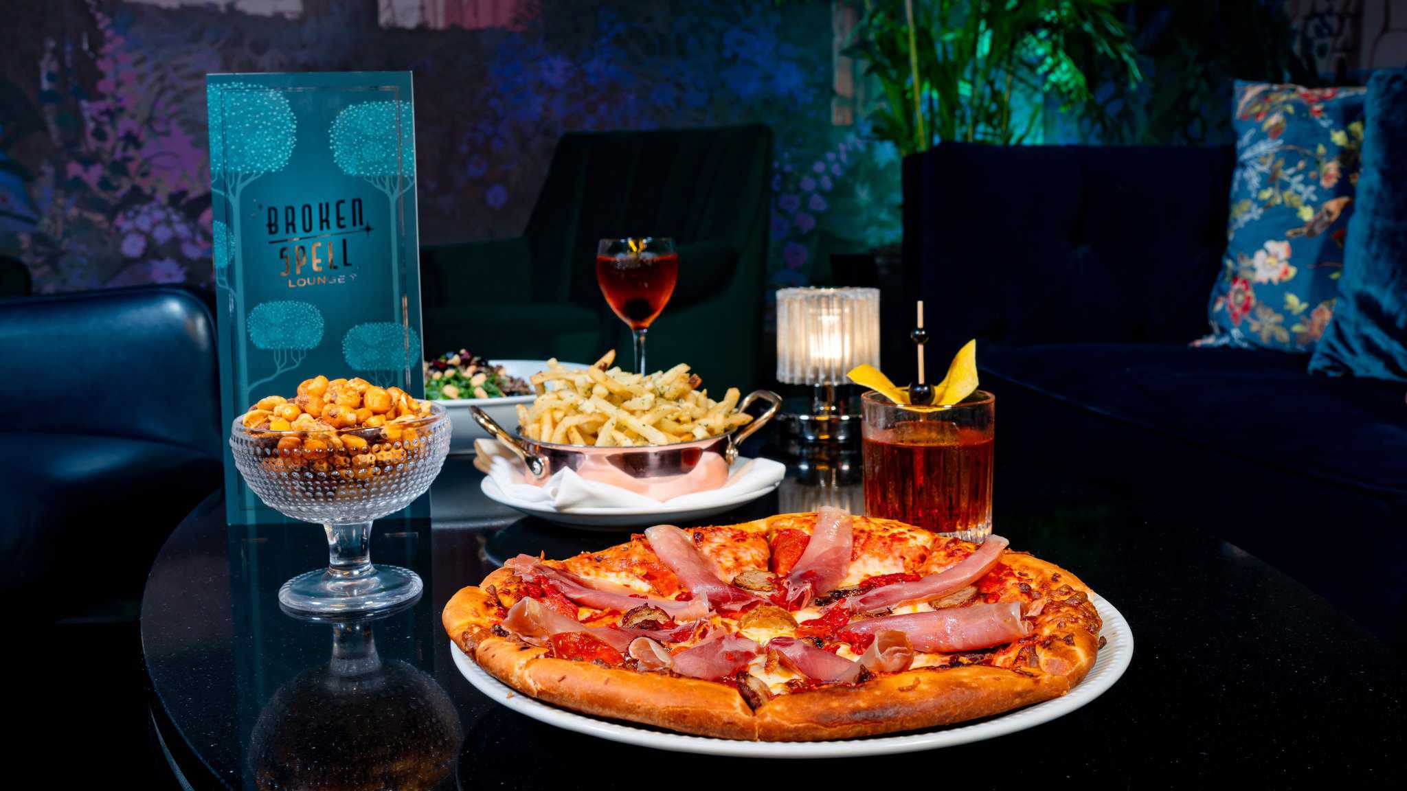 At the enchanting Broken Spell Lounge in Disneyland, a cozy setting awaits: a table featuring a prosciutto-topped pizza, seasoned nuts, fries with dips, a salad, and two cocktails. The inviting ambiance boasts dim lighting and plush seating in the background.