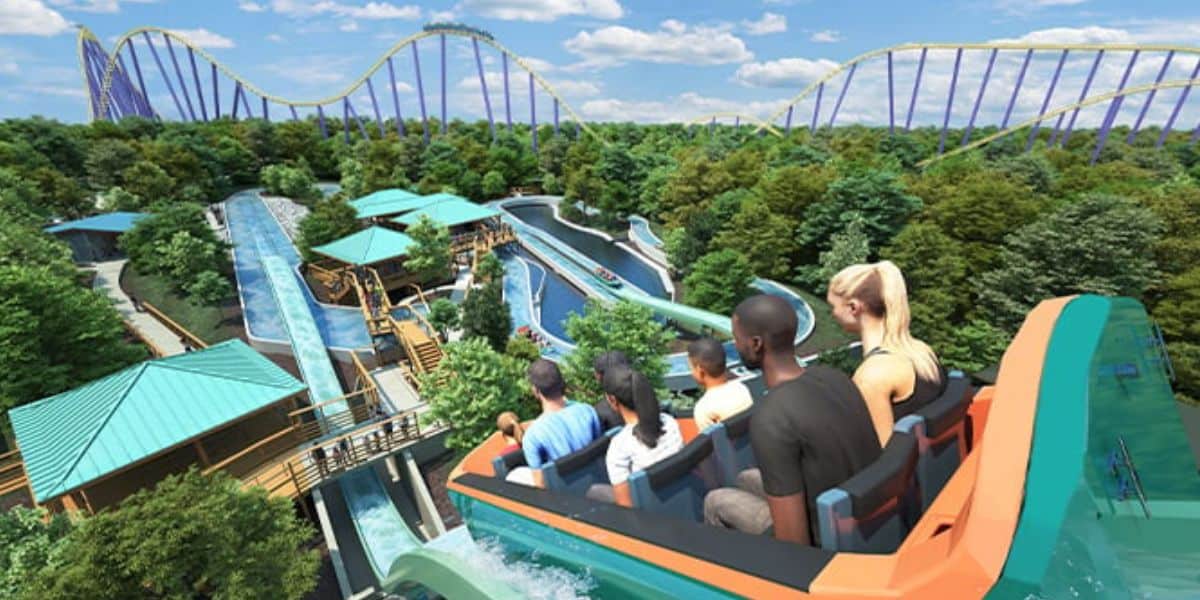 A group of people ride a log flume on a sunny day at SeaWorld San Antonio. The ride descends a steep hill surrounded by lush green trees, while in the background, tall roller coaster tracks rise against blue skies with scattered clouds.