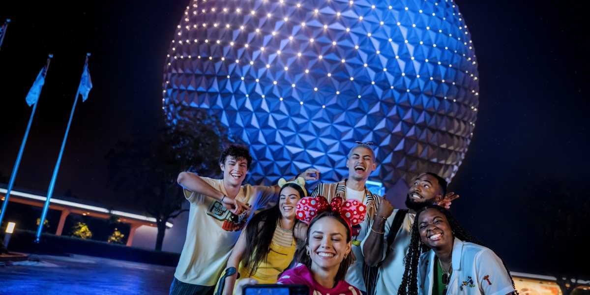 A group of six young adults posing for a selfie at night in front of a large, illuminated geodesic sphere at Epcot during Disney After Hours 2025. The group is smiling and making playful gestures, with one person wearing Minnie Mouse ears. The background is dark with the sphere brightly glowing.