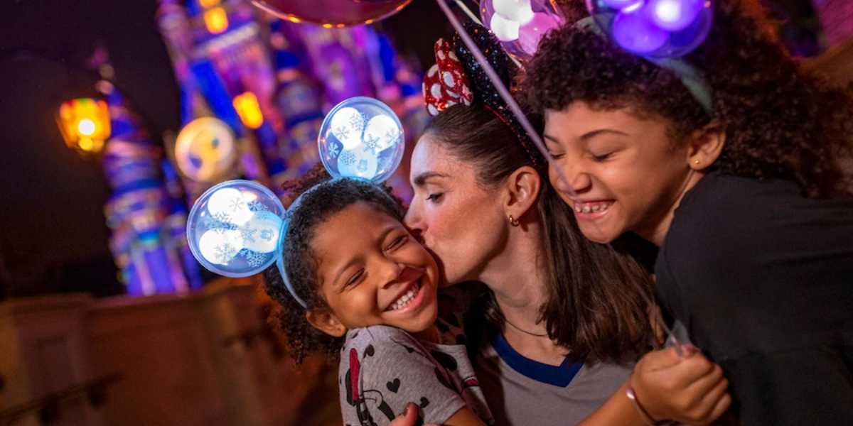 A woman kisses a young child on the cheek while another child smiles and hugs them both. They are all wearing illuminated Mickey Mouse ears, enjoying Disney After Hours at the theme park, with a brightly lit castle in the background.