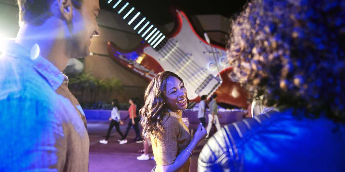 A group of friends enjoying a night out at an amusement park, with a large, illuminated guitar decoration in the background. The people are smiling and laughing, under vibrant lighting that adds a festive atmosphere reminiscent of Disney After Hours events at Walt Disney World.