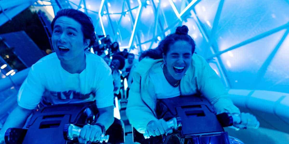 Two people are enjoying a thrilling ride on a roller coaster at Walt Disney World, gripping the handlebars tightly and beaming with excitement. The vibrant, blue lighting around them accentuates the intensity and fun of the Disney After Hours event.