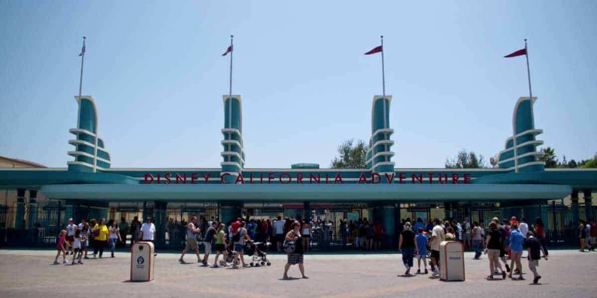 Visitors walk toward the entrance of Disney California Adventure, characterized by its Art Deco-style design, with three flag-topped towers and the park's name prominently displayed above the gate. The scene is busy with families and groups excited to explore the beloved Disney theme park.