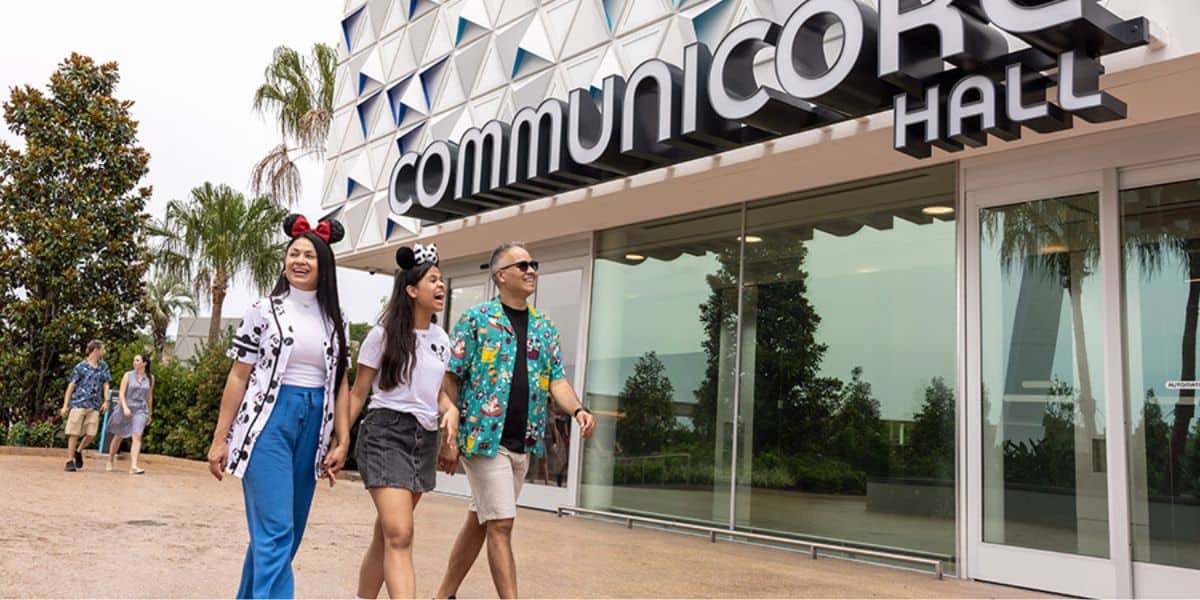 Three people walk side by side, holding hands, in front of CommuniCore Hall. Two are wearing Disney-themed clothing with mouse ears, while one has a colorful shirt. The building has modern architecture with geometric patterns. Trees are visible in the background.