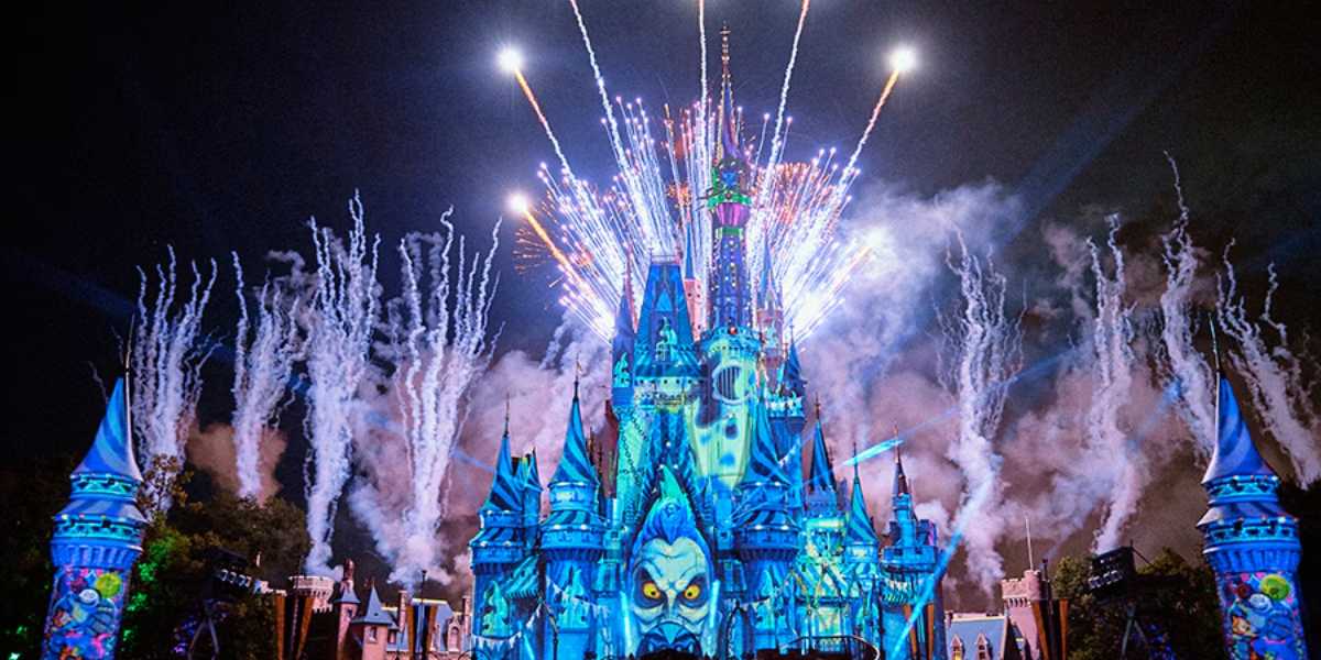 A vibrant fireworks display illuminates the night sky above a brightly lit, fantasy-themed castle adorned with blue and purple hues during the Disney Halloween Party, attended by an excited crowd.