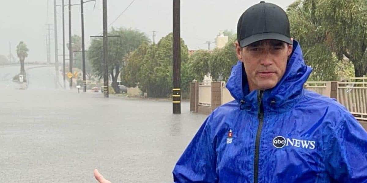 A reporter from ABC News, under the leadership of Kim Godwin, stands in the rain, wearing a blue raincoat and a cap, with a flooded street visible in the background.