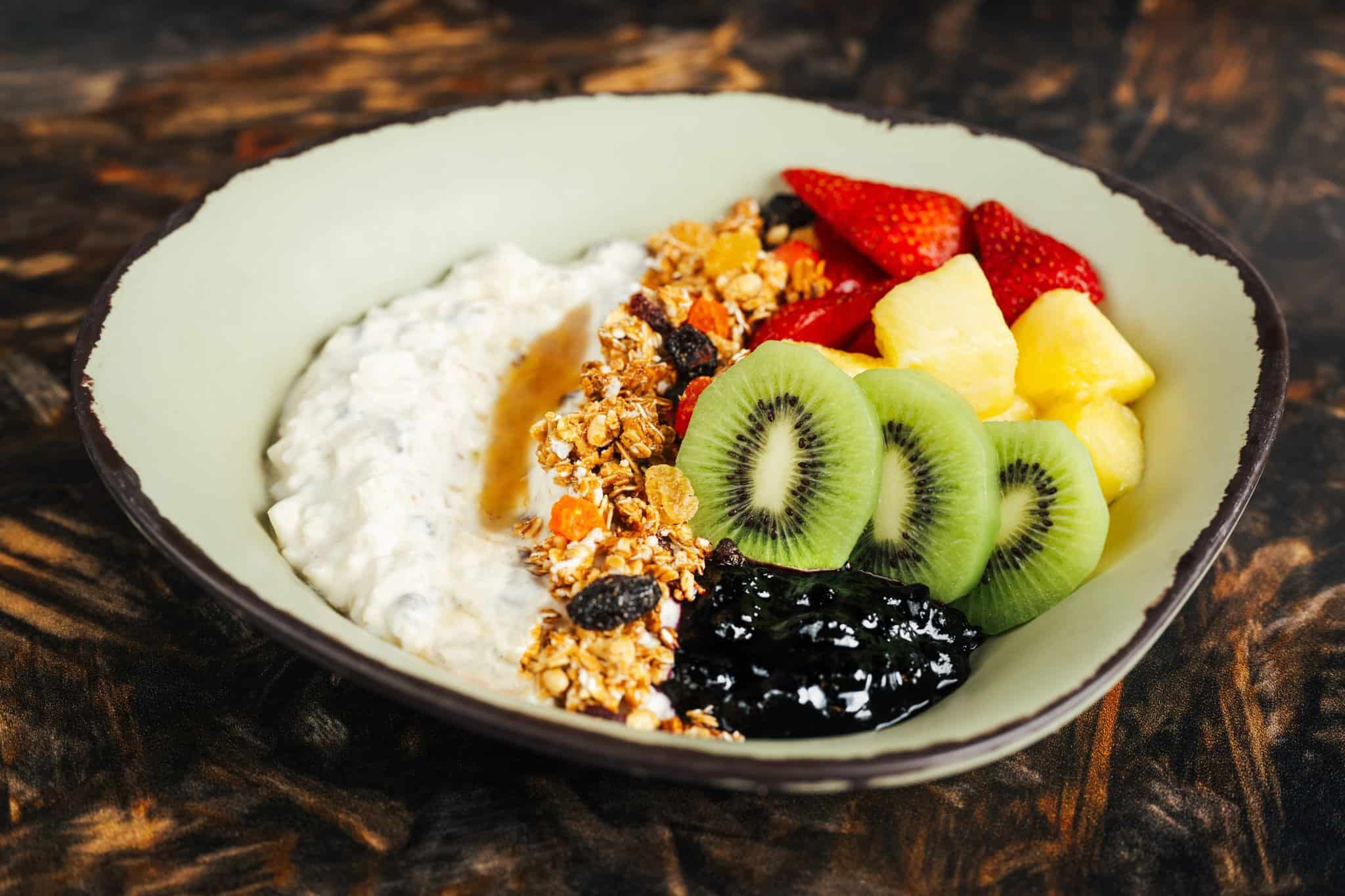 A Satu'li Canteen Breakfast delight: a bowl of yogurt adorned with granola, kiwi slices, pineapple chunks, strawberry halves, and a dollop of dark jam, all elegantly placed on a textured wooden surface.