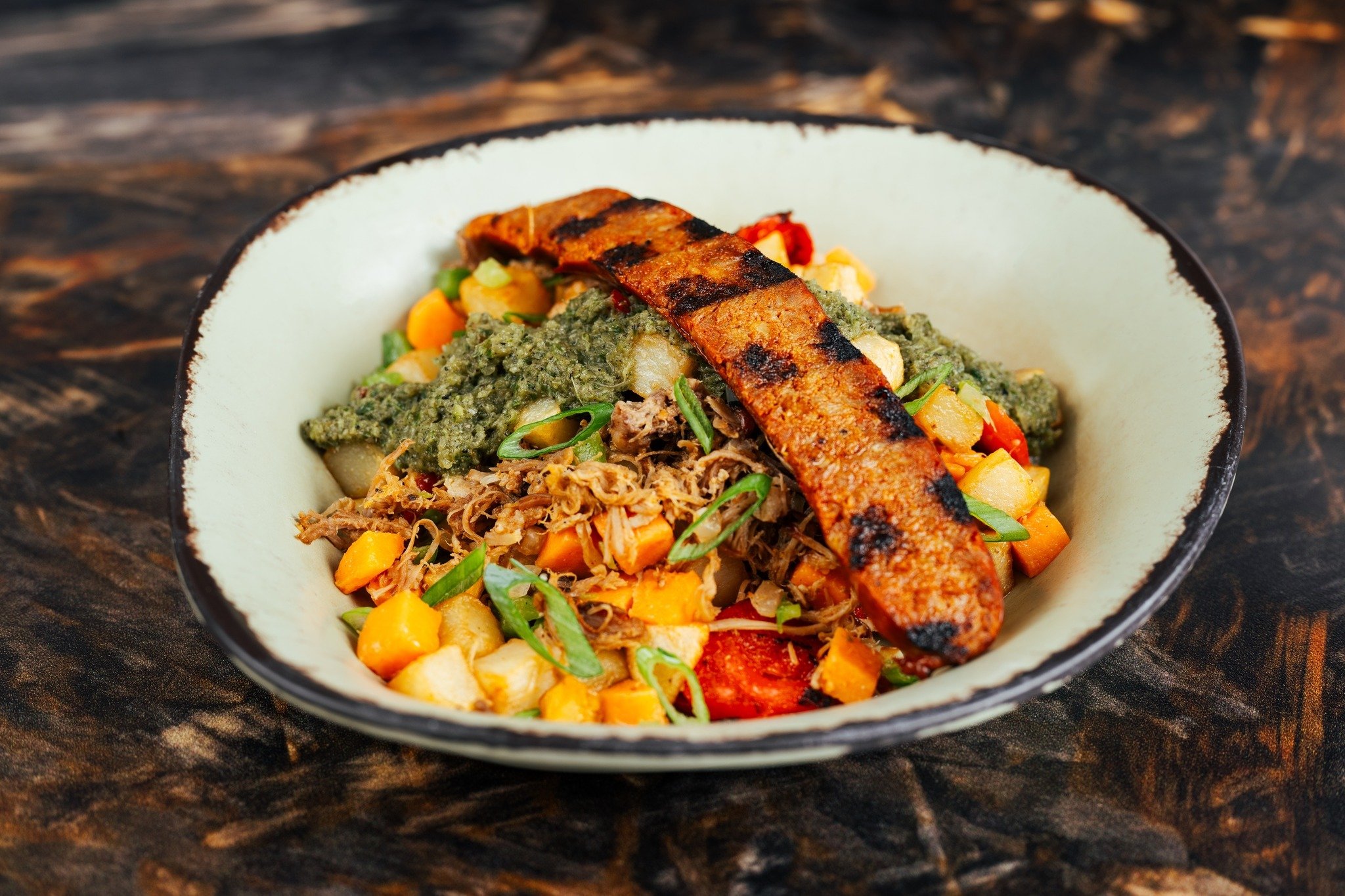 A rustic bowl from Satu'li Canteen Breakfast filled with grilled plantain, shredded pork, roasted vegetables, greens, and a green sauce rests invitingly on a textured wooden surface.