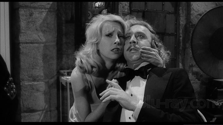 A black and white image depicts Teri Garr and a man in formal attire. The woman, with a worried expression, holds the man's face as he looks upward, appearing concerned. They stand inside a dimly lit room with a gramophone softly playing in the background.