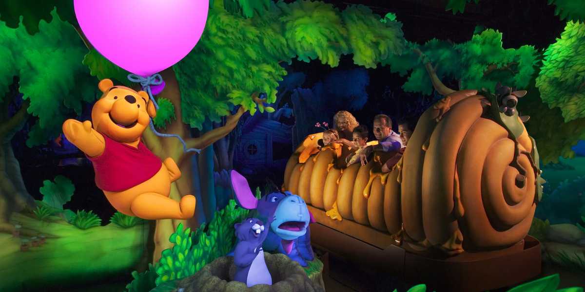 A colorful Disney attraction featuring Winnie the Pooh holding a pink balloon, a snail-shaped vehicle with riders, and various characters from the Hundred Acre Wood, including Eeyore, Piglet, and Tigger, surrounded by green, leafy scenery.