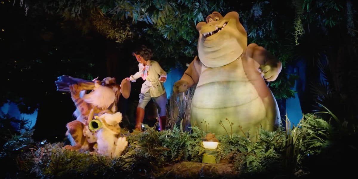 A jungle scene with animatronic characters. A young boy in explorer attire stands next to a large friendly-looking crocodile and a dog wearing goggles, reminiscent of Tiana's Bayou Adventure. They are surrounded by lush greenery and appear to be part of an enchanting theme park attraction.