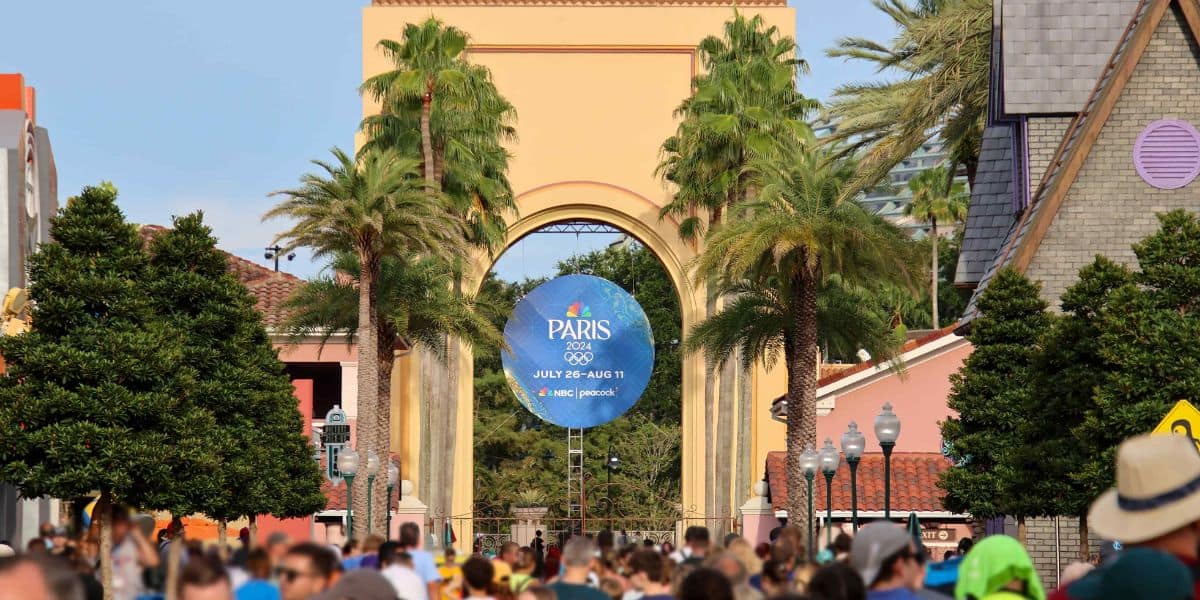 A large plaster archway with an advertisement for the 2024 Paris Olympics greets visitors at Universal Orlando Resort. The promo circle features the Olympic logo, "Paris 2024," and the dates "July 26 - August 11." Dense crowds walk beneath the arch, flanked by palm trees and nearby buildings.