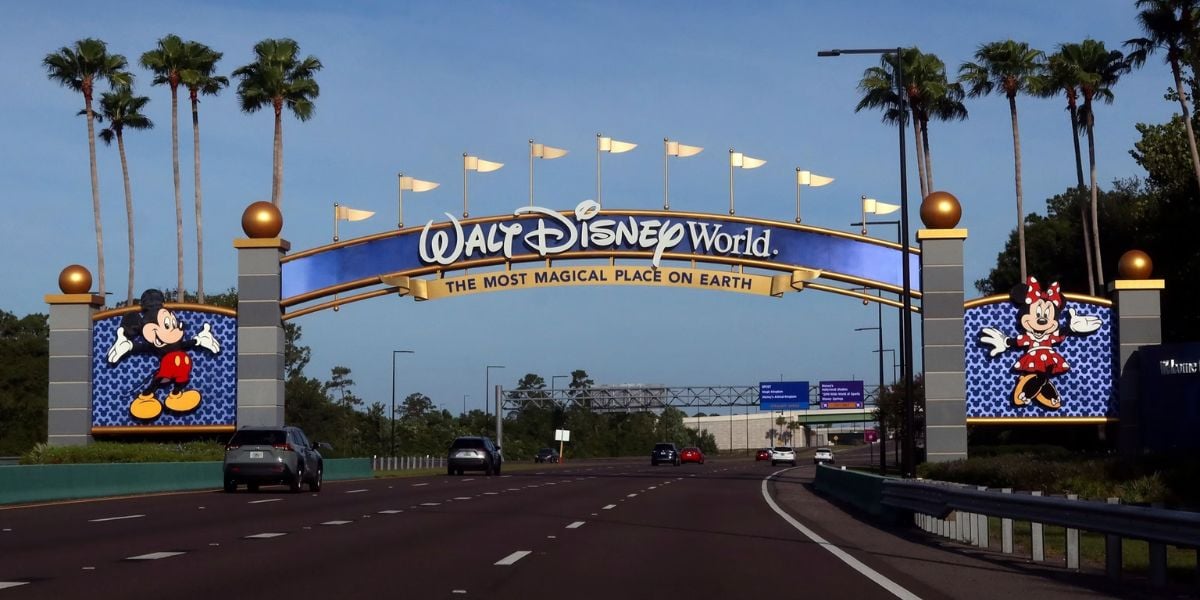 An entrance archway to Walt Disney World with the slogan "The Most Magical Place on Earth." It features images of Mickey Mouse on the left and Minnie Mouse on the right. The road is lined with palm trees under a clear blue sky.