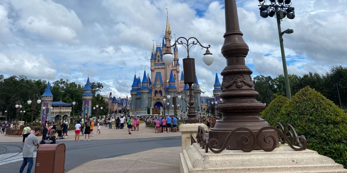 A large Cinderella-style castle at a theme park, surrounded by trees and lamp posts. People are walking and gathering around the area on a cloudy day.