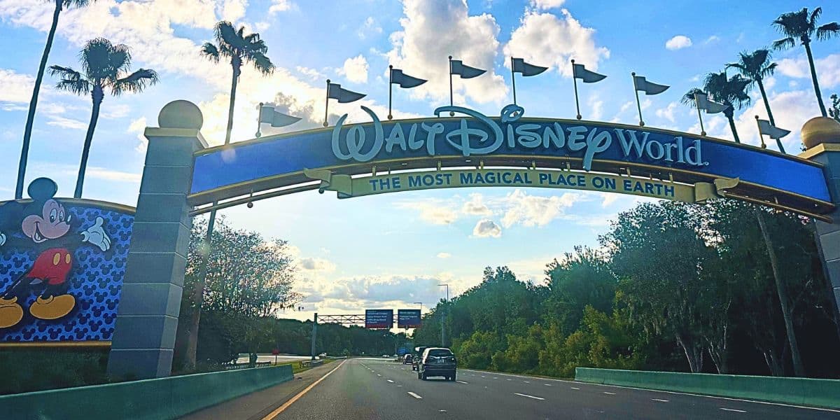 A large, colorful archway sign welcomes visitors to WWDW, featuring its iconic slogan "The Most Magical Place on Earth." The road leading to the entrance is lined with palm trees and includes an image of Mickey Mouse. The sky is partly cloudy. This is the entrance to WDW Resort in Florida.