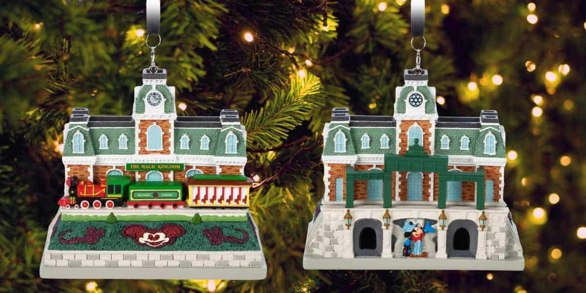 Two park-themed ornaments are shaped like festive buildings adorned with lights and holiday decorations. One ornament features a small train, while the other has a character statue at the entrance. Both Disney releases hang on a lush, illuminated Christmas tree.