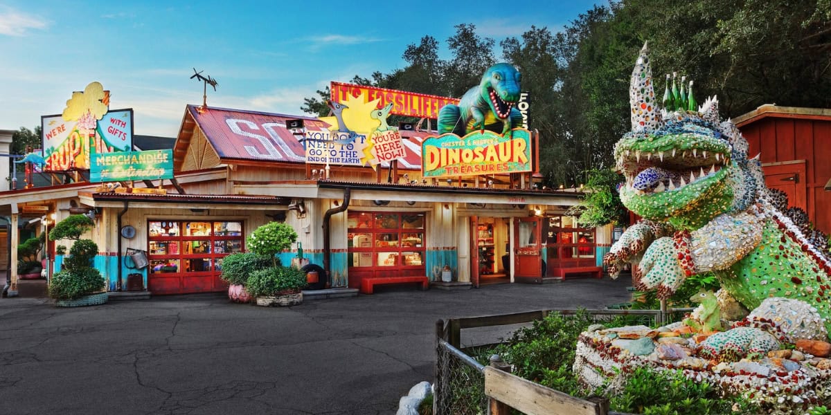 A colorful roadside store adorned with vibrant signs and a dinosaur theme. A large dinosaur sculpture made of various materials stands in the foreground, while another dinosaur sculpture is on the roof. The shop is surrounded by green trees and plants.