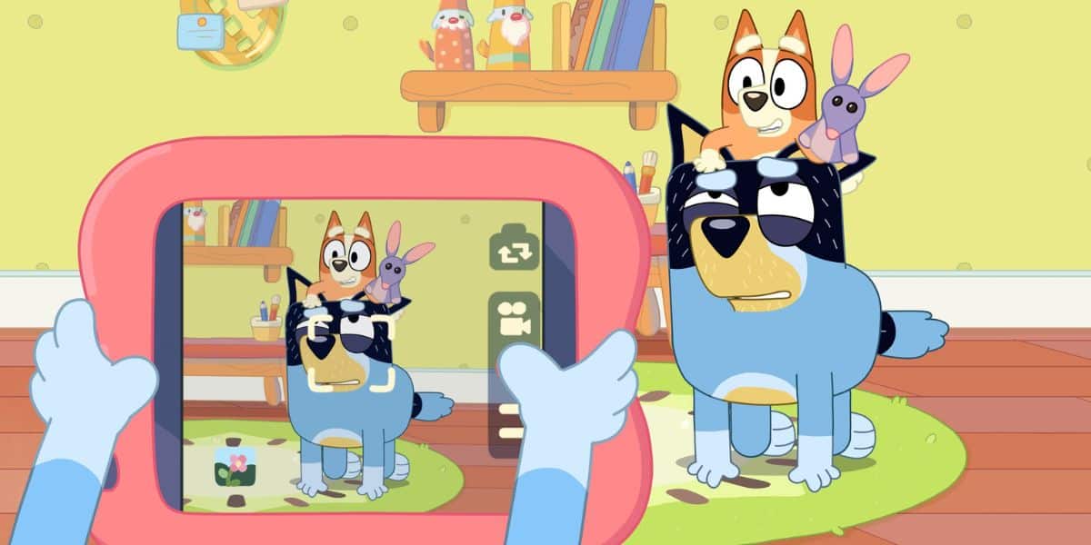 A scene from the animated series "Bluey" shows Bluey taking a photo of her dad, Bandit, who looks tired. Bluey is sitting on Bandit's back, holding a toy bunny, while another toy bunny rests on Bandit's head. The background features toys and a shelf adorned with more beloved items from Bluey's world.