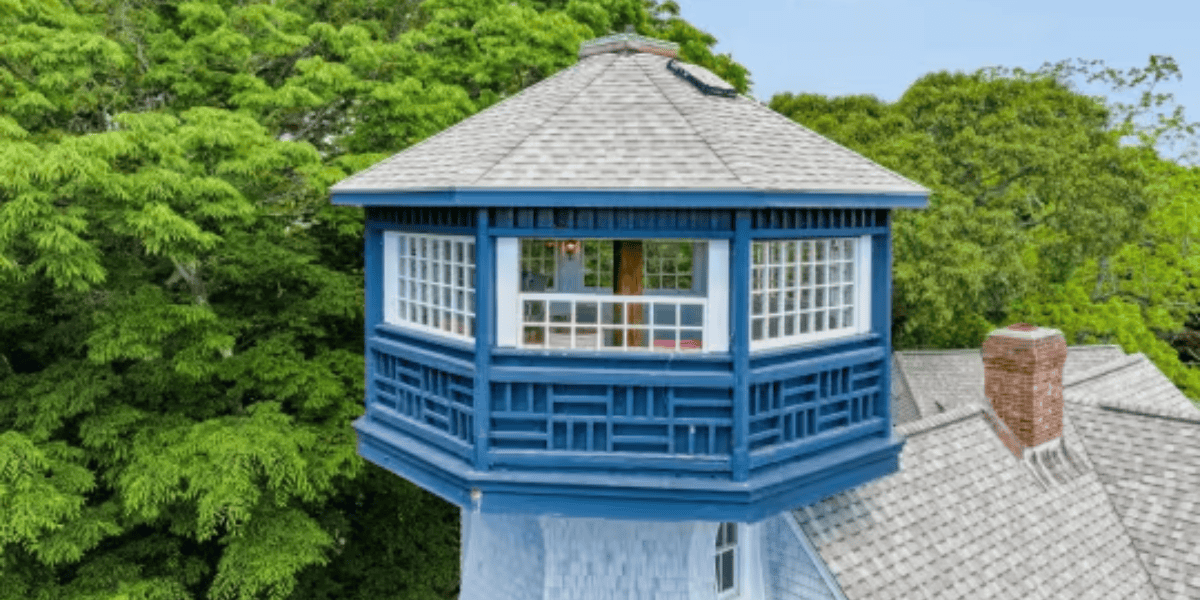 A blue octagonal room perched above a gray-shingled Cape Cod home, where Walt Disney wrote Fantasia, features large windows providing a panoramic view. The house is surrounded by lush green trees, with a small chimney visible on the roof. The scene is under a clear blue sky.