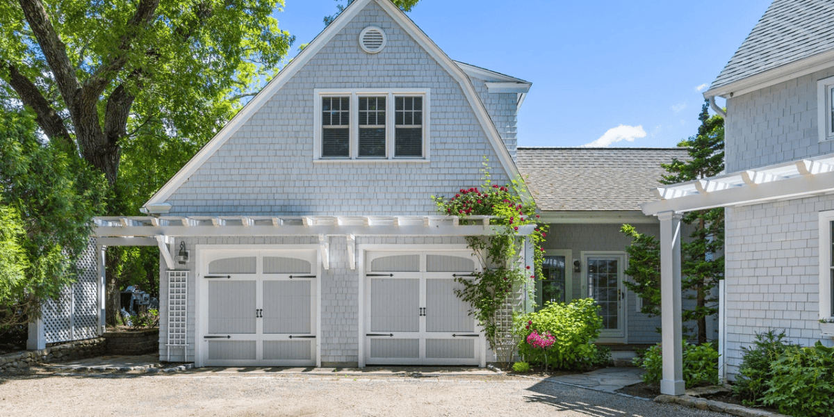 A charming Cape Cod home, reminiscent of where Walt Disney wrote Fantasia, features two large white garage doors adorned with decorative hinges and vine plants. There's an overhang above the garage, and a lush tree and garden flank the house, all enhanced by clear blue skies.