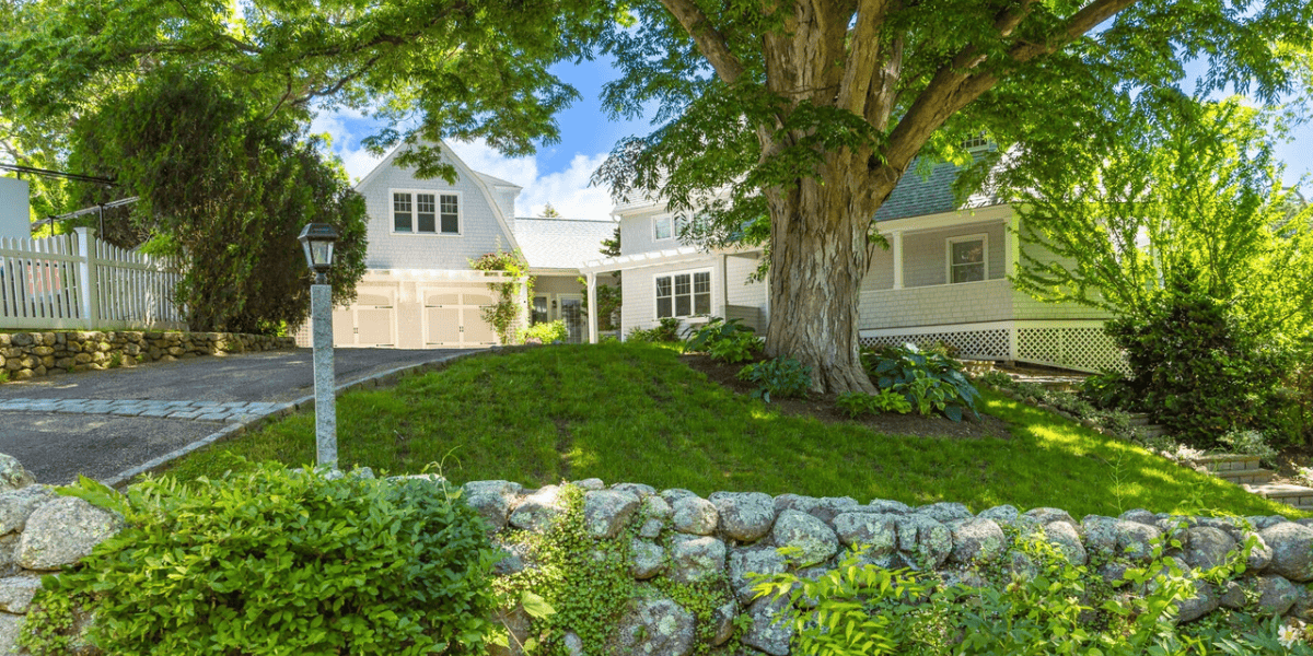 A charming white two-story Cape Cod home, reminiscent of the one where Walt Disney wrote Fantasia, is nestled among lush green trees. The property features a stone retaining wall, a well-maintained lawn, and an inviting pathway leading to the front entrance. The sky is clear with a few scattered clouds.