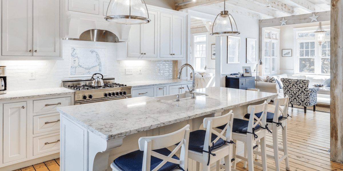 A modern kitchen with white cabinetry, marble countertops, and a central island with four white bar stools sits elegantly in this Cape Cod home where Walt Disney wrote Fantasia. Stainless steel appliances, including a stove with a range hood, are featured. Two pendant lights hang above the island, and a cozy seating area is visible in the background.