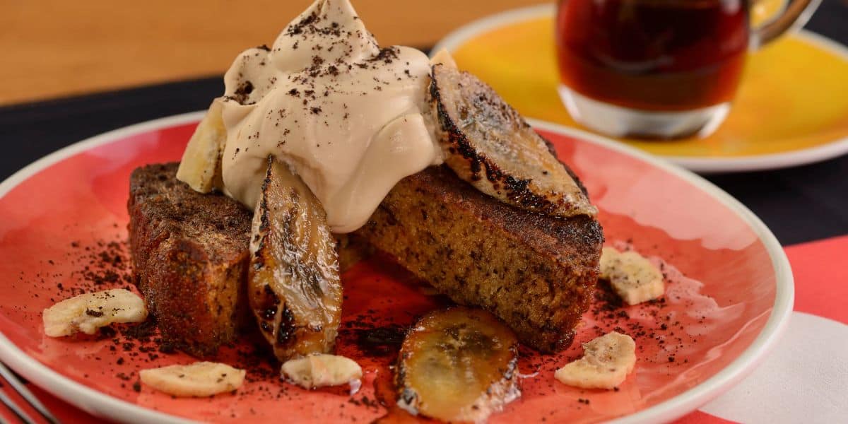 A plate with slices of banana bread topped with caramelized bananas and a dollop of whipped cream. Sprinkles of chocolate or coffee powder are scattered on top. A cup of coffee is in the background.