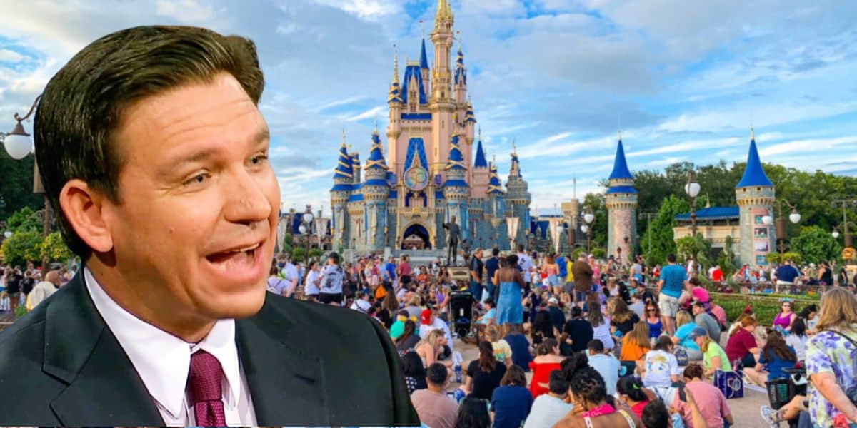 Ron DeSantis, speaking with an open mouth, is superimposed in the foreground of a lively crowd gathering in front of a castle at a Disney theme park. The sky is partly cloudy.