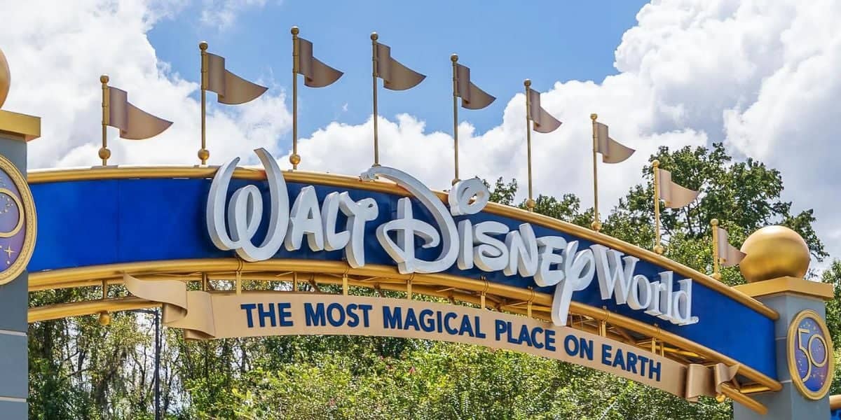 An entrance sign to walt disney world, featuring a large blue banner with the park's name in stylized white lettering, flanked by flags, under a clear blue sky with fluffy clouds.