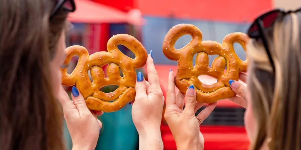 Two people holding pretzels shaped like a cartoon mouse head, showcasing the treats towards the camera. The background is blurred, featuring a mix of red and turquoise colors, suggesting a festive or amusement park setting. The individuals have sunglasses on their heads.