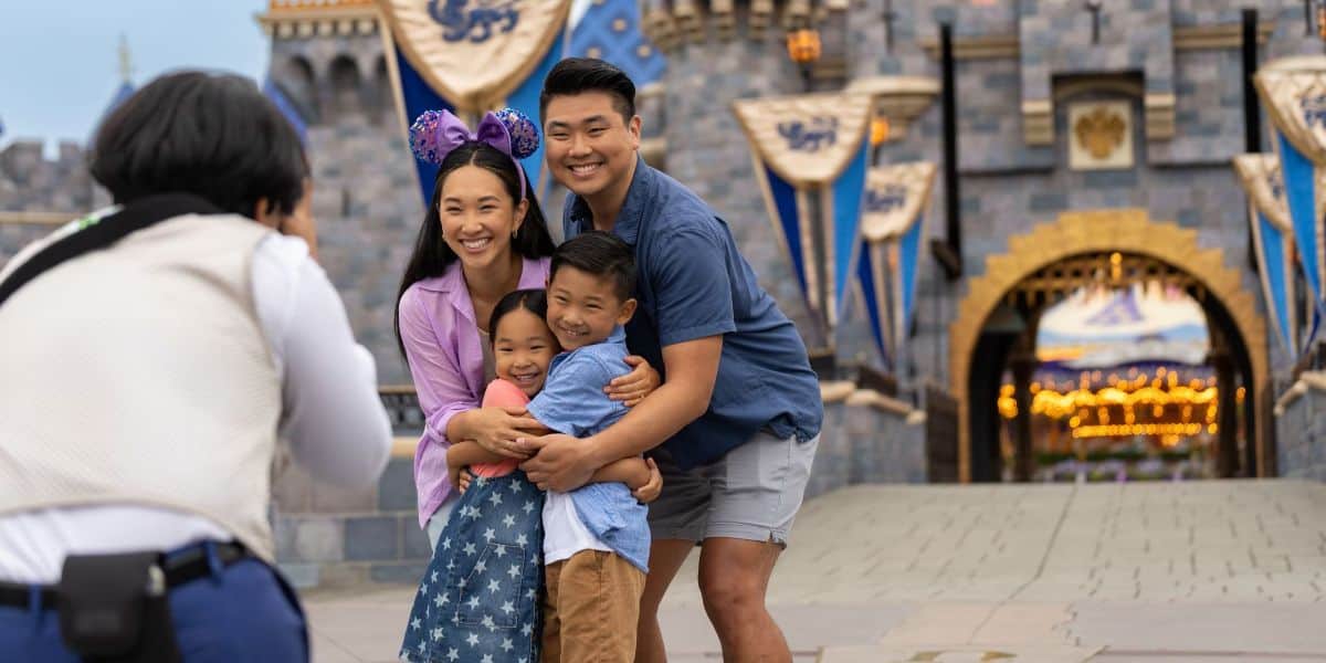 A family of four, consisting of two adults and two children, poses for a photo in front of a castle at a themed amusement park. The woman wears Minnie Mouse ears with a bow, and they all smile happily while hugging each other.