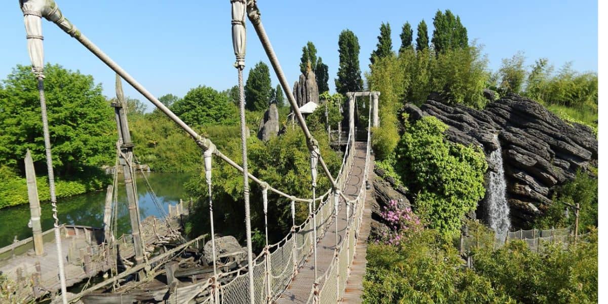 A suspension bridge with wooden planks stretches over a serene river surrounded by lush greenery and rocky formations. Tall trees and vibrant plants border the scene, with bushes and flowers adding color. The sky is clear, enhancing the bright, tranquil atmosphere—much like a hidden gem in Disneyland.