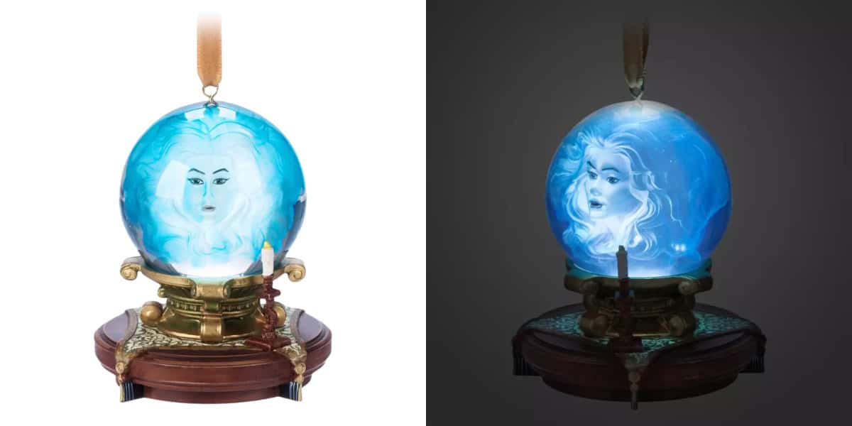 A glowing blue crystal ball featuring Madame Leota's mystical face, surrounded by a golden stand with intricate designs, sits on a wooden base. This enchanting piece, reminiscent of the spooky Disney ornaments collection, showcases one side in light and the other in darkness.