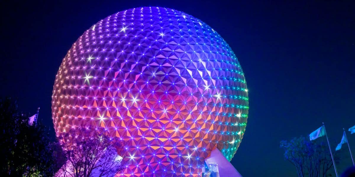 A large geodesic sphere, known as Spaceship Earth, is illuminated in vibrant rainbow colors against the night sky. Flags can be seen in the bottom right near trees silhouetted by the glow of the lights.