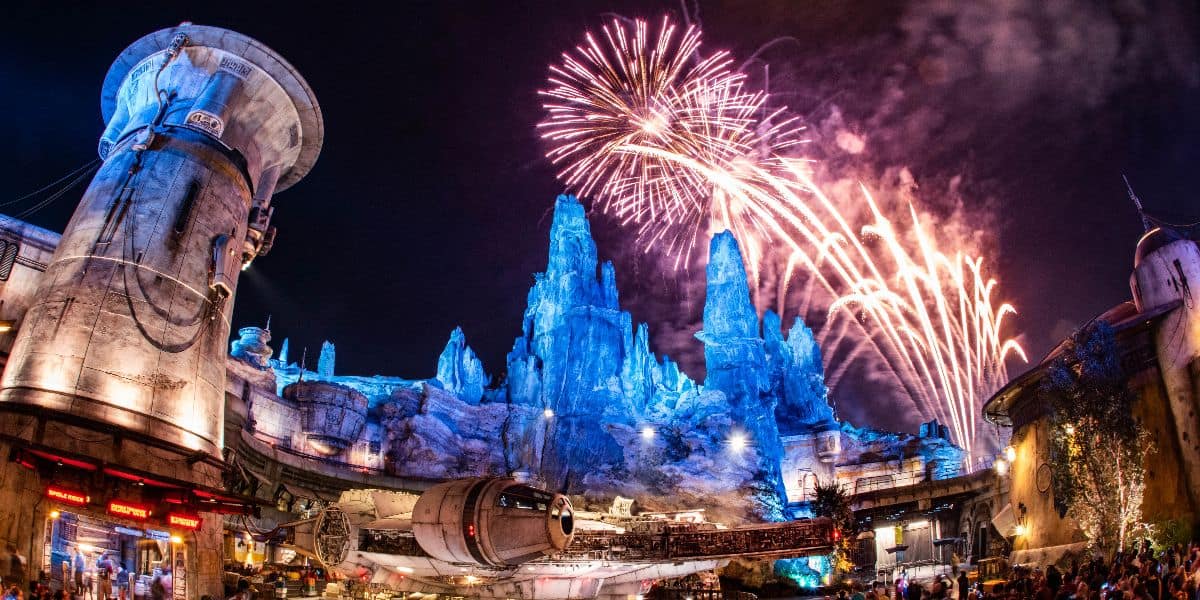 A majestic display of fireworks illuminate the night sky over a bustling Star Wars-themed park, with the iconic Millennium Falcon in the foreground and towering rock formations glowing ethereally in the backdrop.