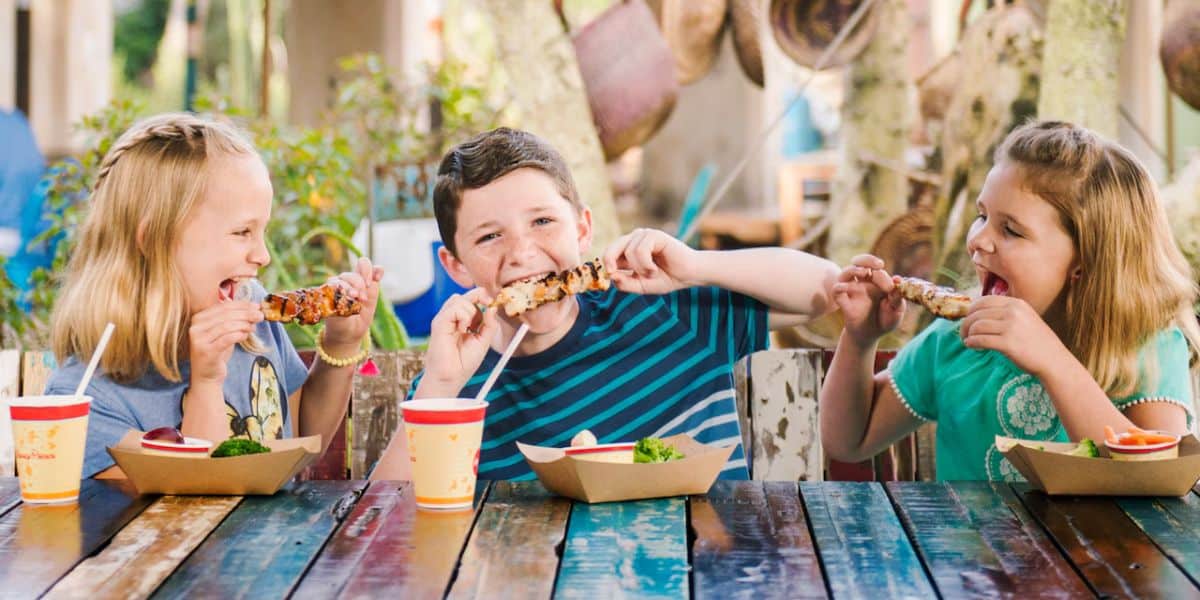 Three children are sitting at a colorful outdoor table at Walt Disney World Resort, enjoying skewers of food. The child on the left wears a yellow dress, the child in the middle wears a blue and black striped shirt, and the child on the right wears a turquoise dress. They are all laughing and having fun.